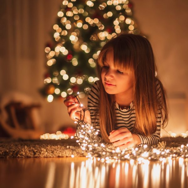 Beautiful little girl lying on the floor next to nicely decorated Christmas tree, arranging Christmas lights and smiling